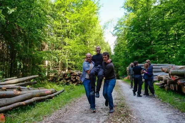Drei Personen tragen einen Mann auf einem Waldweg zwischen Holzstapeln